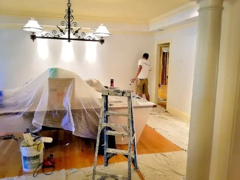 painter cuting-ing the wall with a brush ahead of rolling the walls. Furniture is covered with plastic sheeting and floors with drop cloths