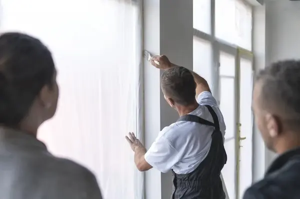 Painter performing final touch-ups on a wall near a window while a satisfied couple looks on in the background.